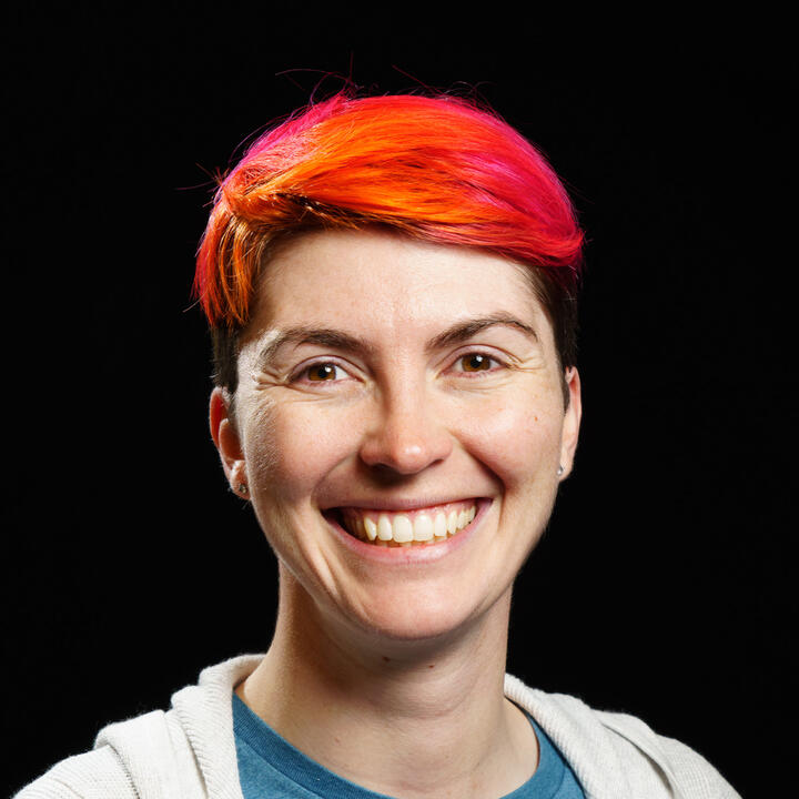 a white woman with pink and orange hair, wearing a gray sweater over a blue shirt